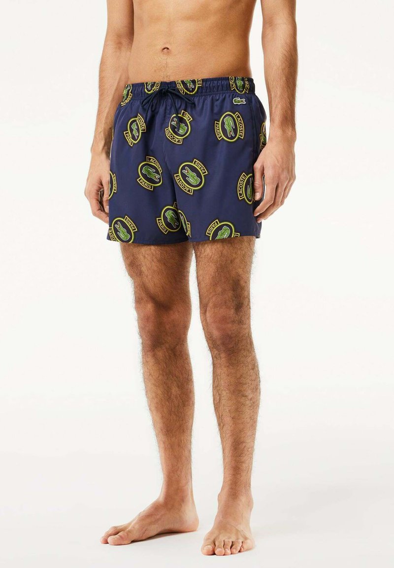 Bañadores de natación en color navy con cintura elástica y cordón, que presentan un logo de cocodrilo verde repetido y acentos de texto en forma circular. Textura suave.