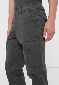 Pantaloni de cargo gri, dintr-un material texturat, cu multiple buzunare laterale și închidere cu nasture, având o croială lejeră și detalii cusute.