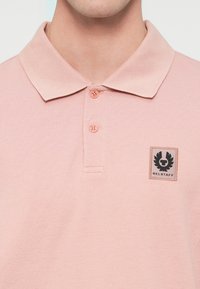 Homme portant un polo rose clair avec deux boutons et un écusson du logo Belstaff sur le côté gauche de la poitrine.