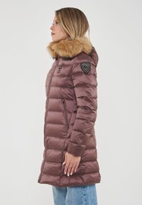 Donna di profilo a sinistra che indossa un lungo giubbotto imbottito mauve con cappuccio foderato in pelliccia e tasche laterali, abbinato a jeans azzurri chiari.