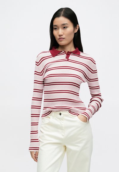 Polo manches longues en tissu côtelé à rayures rouges et blanches. Il présente un col rouge et des boutons assortis, porté avec un pantalon écru.