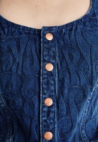 Top in denim con un motivo stampato blu scuro, caratterizzato da bottoni a pressione color rame sul fronte e un collo rotondo.