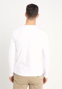 Bílé tričko s dlouhým rukávem a kulatým výstřihem, vyrobené z měkkého materiálu, zobrazené zezadu, v kombinaci se světle zbarvenými kalhotami.