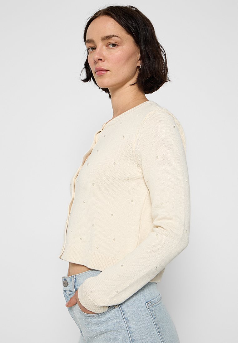 Gestrickte cremefarbene Strickjacke mit perlartigen Verzierungen, langen Ärmeln, cropped Design und rundem Ausschnitt. Weicher Griff.