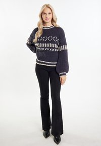 Marine gebreide trui met crème geometrische patronen en puffed mouwen, gecombineerd met zwarte flared jeans en puntige zwarte schoenen.