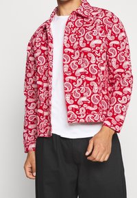 Mann trägt eine rote Jacke mit Paisley-Muster über einem weißen Hemd und schwarzen Hosen, steht vor einem schlichten hellen Hintergrund.