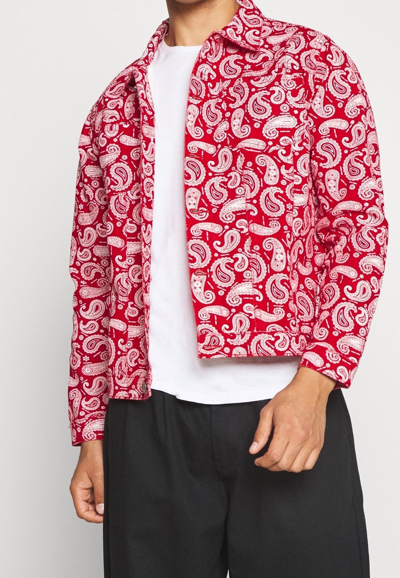 Mann trägt eine rote Jacke mit Paisley-Muster über einem weißen Hemd und schwarzen Hosen, steht vor einem schlichten hellen Hintergrund.