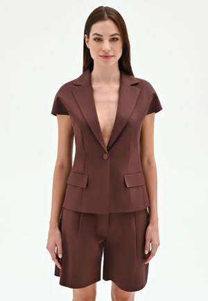 Femme portant un blazer marron à manches courtes avec un profond décolleté en V et un short assorti arrivant aux genoux, debout devant un fond blanc uni.