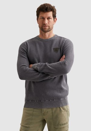 Muž so založenými rukami, oblečený v štruktúrovanom sivom svetri a olivových cargo nohaviciach, stojaci pred jednofarebným svetlým pozadím.