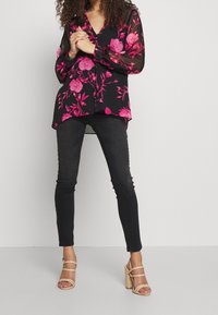 Blusa nera trasparente con grande motivo floreale rosa, maniche lunghe e scollatura a V. Abbinata a jeans skinny neri e sandali con tacco quadrato beige.