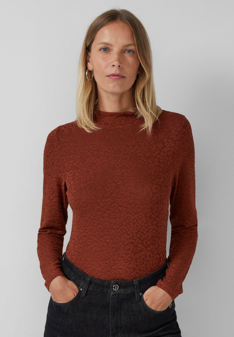 Femme aux cheveux blonds raides portant un haut à motifs à manches longues couleur rouille et un pantalon noir, se tenant avec les mains dans les poches devant un fond gris.