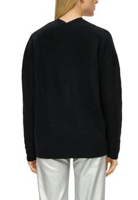 Schwarzer strukturierter Strickpullover mit V-Rücken-Design, ausgestattet mit überschnittenen Schultern und einem lässigen Schnitt, kombiniert mit glänzenden silbernen Hosen.