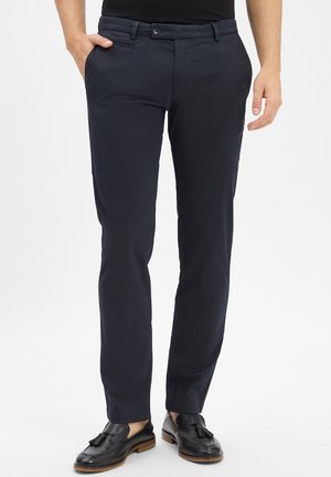 Mann in schmal geschnittenen, dunkelblauen Hosen mit Seitentaschen und schwarzen Leder-Tassel-Loafers vor weißem Hintergrund.