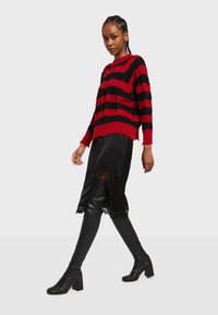 Červeno-čierny pruhovaný pletený sveter s potrhanými okrajmi, kombinovaný s čiernou saténovou sukňou s čipkovými akcentami a čiernymi čižmami na vysokom podpätku po kolená.