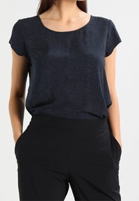 Modrý top s krátkým rukávem a jemným tečkovaným vzorem, kulatým výstřihem a splývavou látkou, v kombinaci s černými kalhotami s vysokým pasem. Nejasný pozadí.