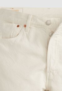 Gros plan sur un jean en denim blanc cassé montrant la ceinture, la fermeture à bouton métallique, la boucle de ceinture et la poche avant avec des rivets en cuivre.