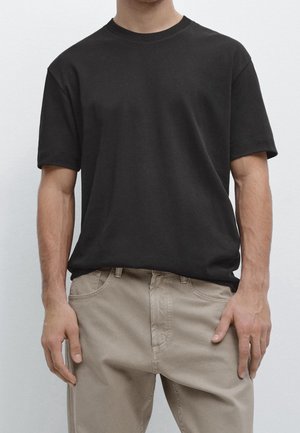 Homme portant un t-shirt noir uni à manches courtes légèrement rentré dans un pantalon beige, debout devant un fond clair uni.