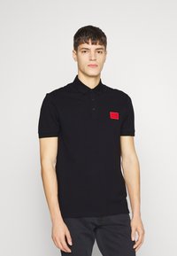 Polo shirt noir avec un col, des manches courtes et un patch de logo rouge sur la poitrine gauche. Le tissu semble lisse et ajusté.