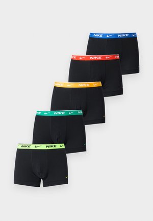 Mužský model stojaci na koberci, oblečený v čiernych boxer nohavičkách Nike so zeleným pásom, jedna ruka na bokoch, neutrálny výraz tváre, jednoliaté svetlé pozadie.