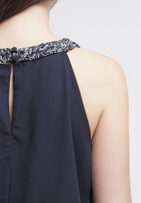 Marinblå halterneck med en pärlbesmyckad halsringning, med en nyckelhålsöppning bak och mjuk, flödande struktur.