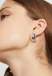 Pendientes de metal plateado con un diseño geométrico que presenta formas onduladas e entrelazadas. La textura suave y el acabado pulido realzan el atractivo visual.