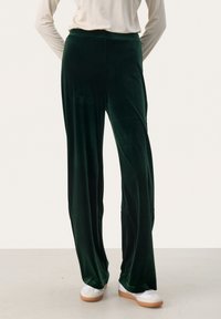 Pantaloni ampia in velluto verde scuro, con una texture liscia e una cintura elastica, abbinati a sneakers bianche per un contrasto.
