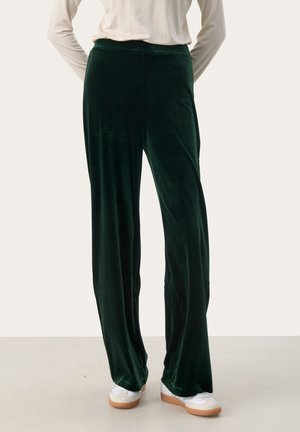 Fluwelen wijde broekspijpen in diep groen, met een gladde textuur en een elastische tailleband, gecombineerd met witte sneakers voor contrast.