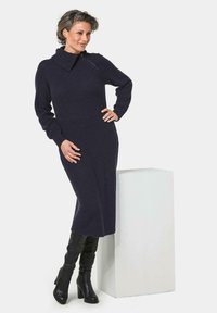 Navy gestricktes Kleid mit hohem Kragen und Reißverschlussdetails, langen Ärmeln und gerader Silhouette. Kombiniert mit knielangen schwarzen Lederstiefeln.