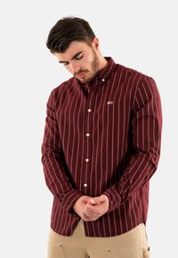Camicia bordeaux a maniche lunghe con righe verticali bianche, colletto button-down e piccolo logo sul petto. Tessuto di cotone con una consistenza liscia.