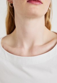 Bílá bavlněná košile s širokým výstřihem, která ukazuje hladkou texturu látky. Viditelné stříbrné kruhové náušnice, zdůrazňující jednoduché designové detaily.