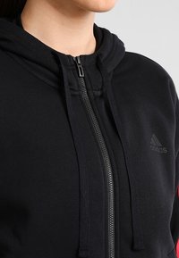 Sweat à capuche noir zippé avec une texture de tissu lisse, des cordons de serrage ajustables et un petit logo embossé sur la poitrine.