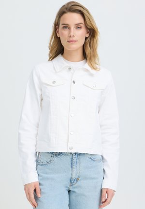 Mujer con cabello rubio hasta los hombros, vistiendo una chaqueta blanca abotonada y jeans azul claro, de pie contra un fondo claro y liso.