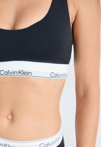 Primo piano del torso di una persona che indossa un reggiseno sportivo nero di Calvin Klein e biancheria intima coordinata con bande del logo visibili.
