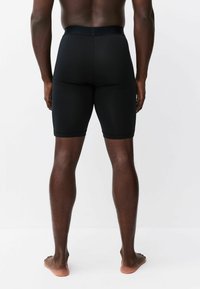 Man standing barefoot facing away wearing fitted black athletic shorts against a plain white background.