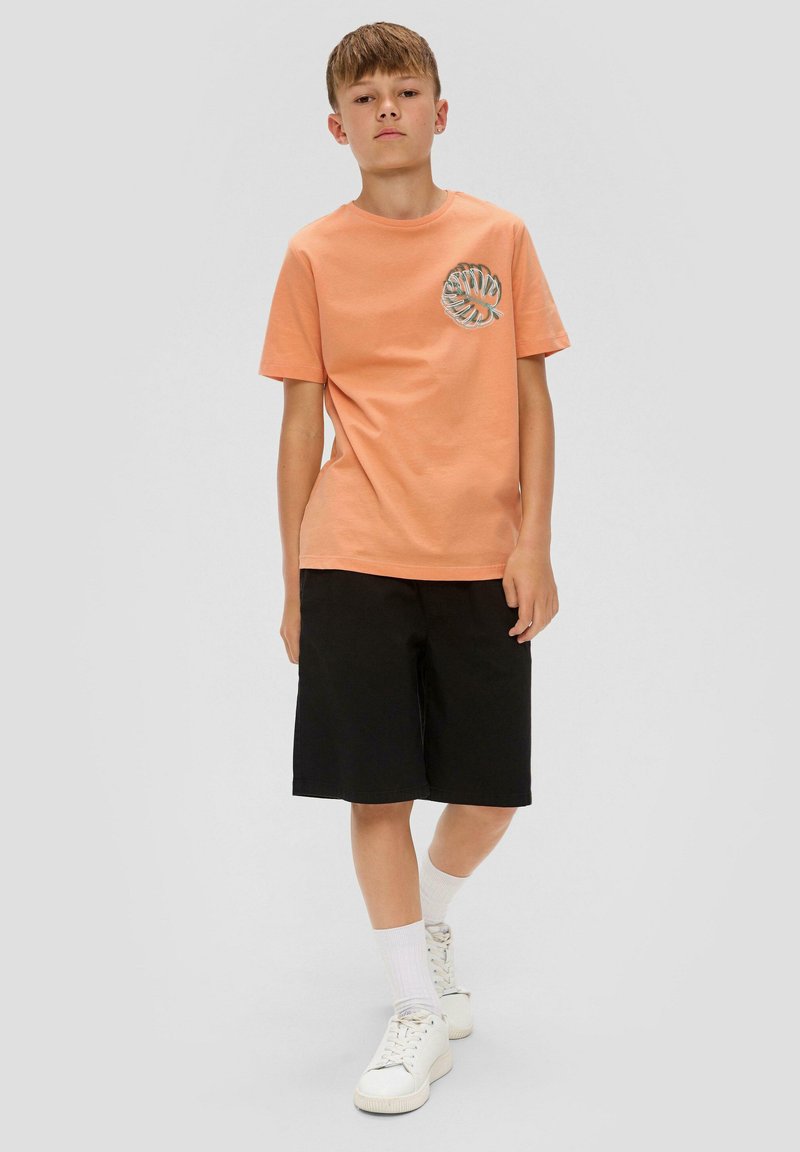 Pomarańczowa bawełniana koszulka z grafiką liścia na klatce piersiowej, w połączeniu z czarnymi szortami. Model nosi białe skarpetki i trampki.