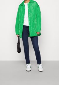 Abrigo acolchado verde con botones a presión, combinado con una camiseta blanca, jeans oscuros, bolso negro y zapatillas blancas con acentos verdes.