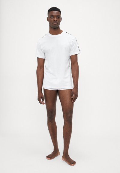 Vitt kortärmat t-shirt i bomull, med en enkel design och minimala accenter samt rundad halsringning. Modellen står barfota.
