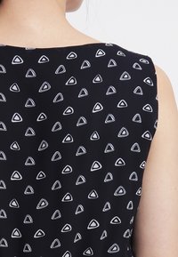 Svart tyg med ett triangulärt vitt mönster, med en slät textur och ärmlös design. Fokusera på bakvy med detalj på halsringningen.