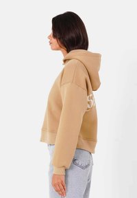 Sudadera recortada de algodón beige con capucha ajustable, puños y dobladillo acanalados. Presenta un detalle del logo en blanco en la parte posterior. Combinada con jeans de color azul claro.