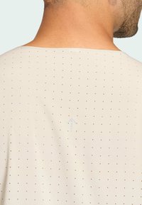 Ανοιχτό μπεζ πουκάμισο με μικρές καφέ κουκίδες, διαθέτει στρογγυλή λαιμόκοψη και διακριτό μπλε σχέδιο βέλους στο πίσω μέρος. Μαλακή υφή υφάσματος.