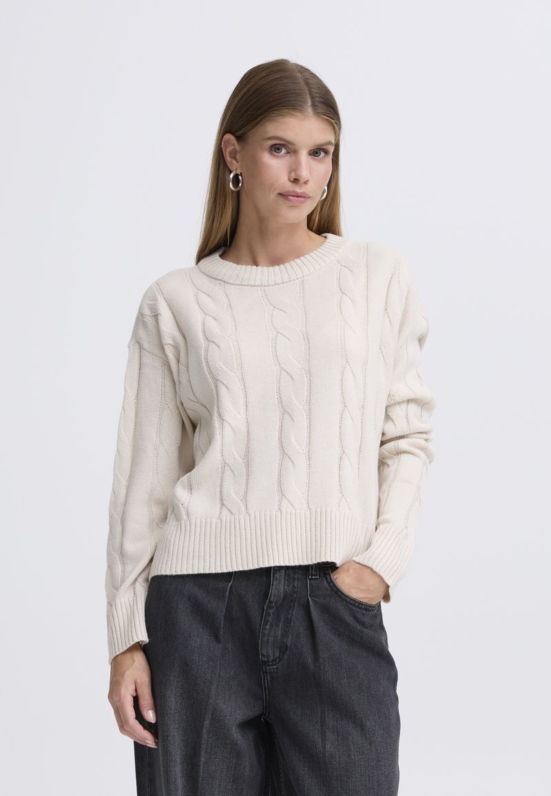 Krem pulover iz kabelsko pletenega blaga z rebrastim izrezom in robom, dolgimi rokavi in sproščenim krojem, kombiniran s temnimi širokimi hlačami.