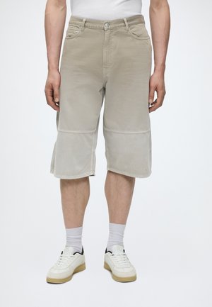 Hombre con pantalones cortos beige claro hasta la rodilla, calcetines blancos, zapatillas blancas con suelas beige y una camisa blanca visible en la cintura.