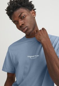 Camiseta de algodón azul claro con mangas cortas y escote redondo. Presenta un logotipo de texto blanco "Originals studio" en el frente.