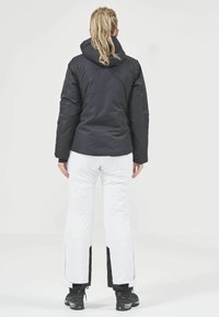 Schwarze isolierte Jacke mit Kapuze, kombiniert mit weißen Schneehosen mit schwarzen Seitenpaneelen und schwarzen Wanderschuhen. Einfaches und funktionales Design.
