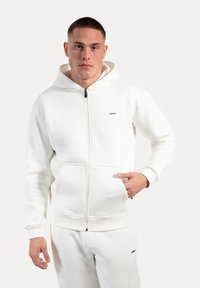 Felpa bianca con zip presenta un cappuccio foderato, tasche frontali, polsini a coste e un piccolo logo sul petto. Il tessuto sembra morbido e testurizzato.