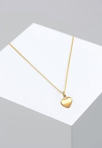 Aranyszínű szív alakú medál nyaklánc, sima, tükröződő felülettel, finom lánchoz rögzítve, geometriai talapzaton kiállítva.