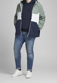 Giacca leggera color block in verde, bianco e blu navy, indossata sopra un hoodie blu navy, abbinata a jeans blu e sneakers bianche.