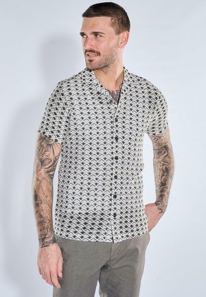 Mann mit Tattoos, der ein kurzärmeliges schwarz-weiß gemustertes Hemd und graue Hose trägt und mit der Hand in der Tasche nach links schaut.