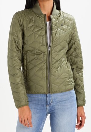 Femme portant une veste bomber matelassée verte avec fermeture éclair devant, col côtelé et poches, associée à un jean bleu clair.