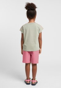 Maglietta verde in cotone a maniche corte, abbinata a pantaloni corti rosa. I sandali presentano una fascia multicolore e una suola scura.